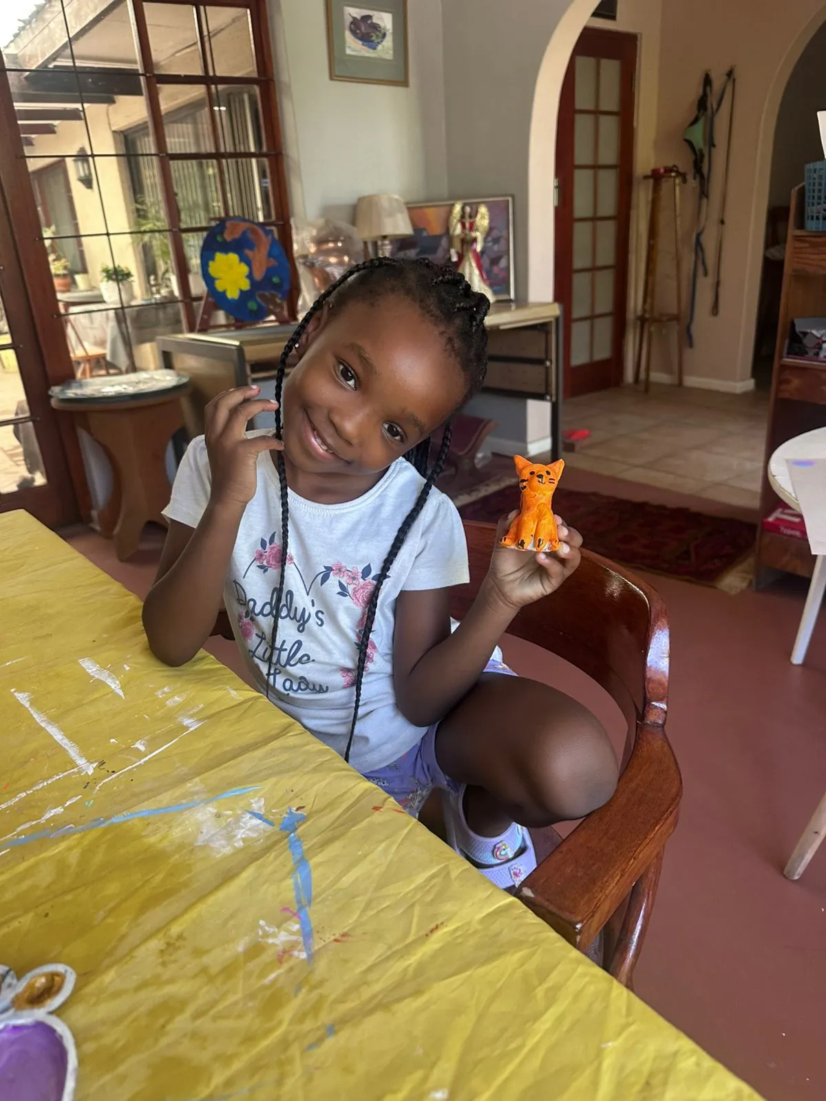 A smiling young girl at a class table proudly holding a small painted clay figurine she made