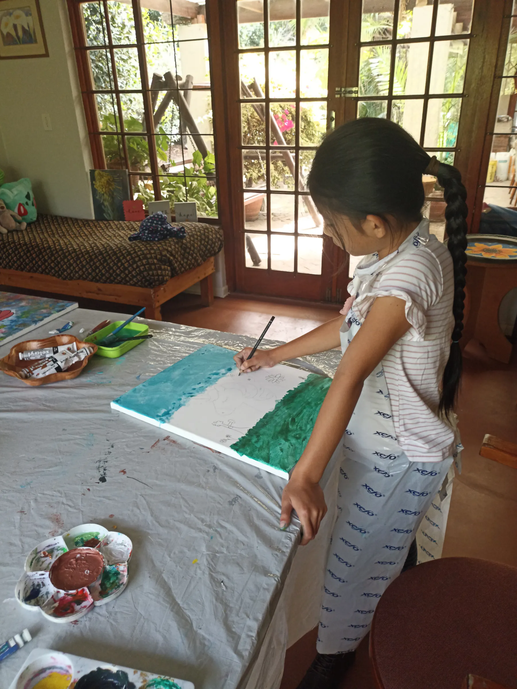 A child painting a green-and-white canvas at a table beside sunlit garden doors