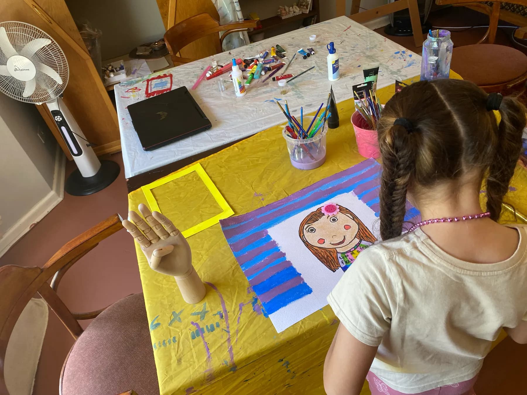 A girl with braids painting a portrait of a child on bright blue and yellow paper at a table