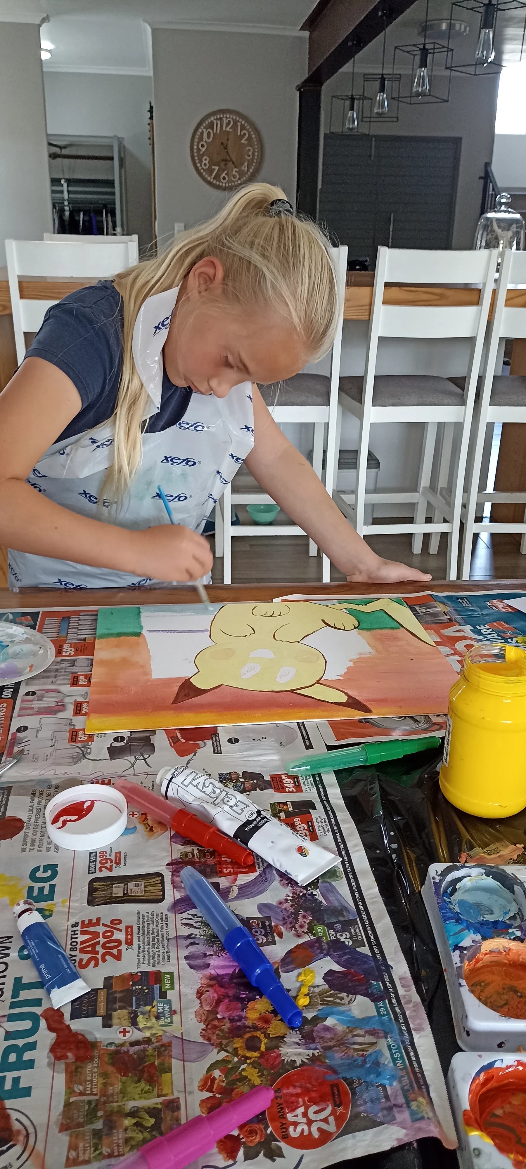 A young girl carefully painting a colourful artwork at a craft-covered table in art class